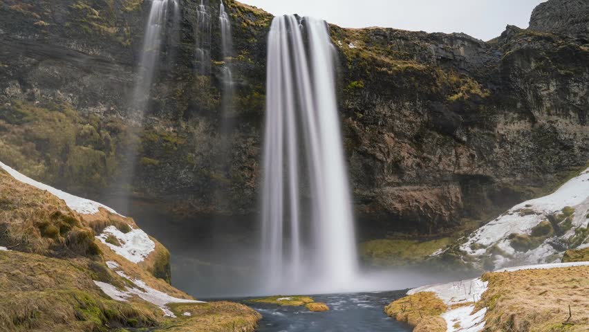 Time lapse of a tall waterfall plunging down a rocky cliff with snow patches.