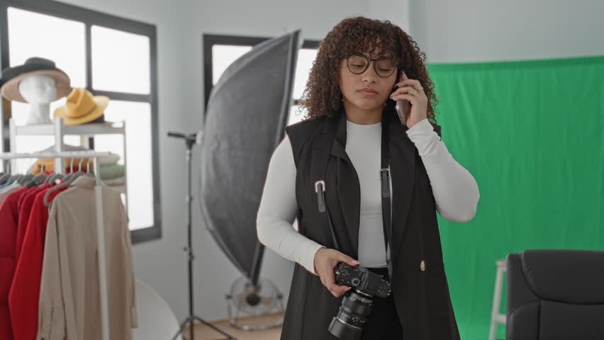 Woman holding dslr camera in one hand and speaking on smartphone, checking settings in studio; concentration planning.