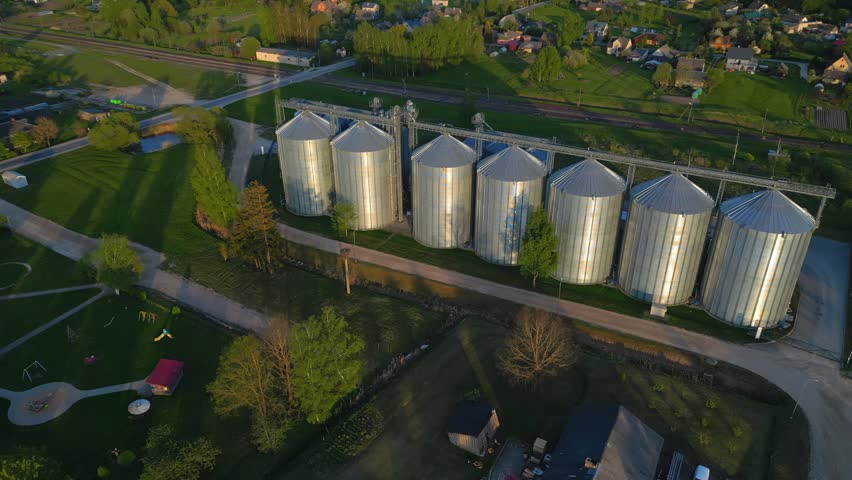 Aerial view of metal grain silos standing in a rural landscape with green fields, trees, and nearby houses in Latvia.