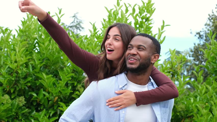 Young caucasian woman getting a piggyback ride from her african american boyfriend, both smiling and looking around happily while spending a lovely day together in a lush green park