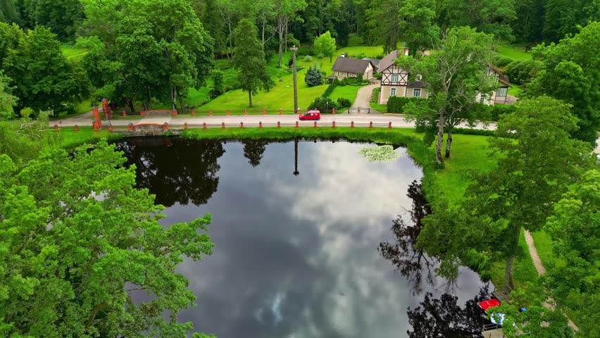 Aerial view of a pond reflecting the sky, with a red car driving on a road past a charming half-timbered house surrounded by lush green trees in Latvia.