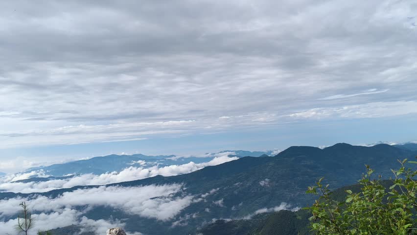 A beautiful high-altitude mountain landscape featuring lush green pine trees and foliage in the foreground. In the background, layers of blue mountain ridges stretch across the horizon beneath a dramatic, overcast sky. Soft white clouds rest in the valleys and slowly roll across the peaks, capturing the serene atmosphere and fresh wilderness of high mountain ranges.
