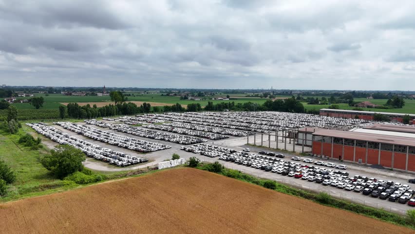 Thousands of parked passenger cars fill a large vehicle distribution center surrounded by agricultural fields, highlighting automotive logistics, inventory management, drone flyover