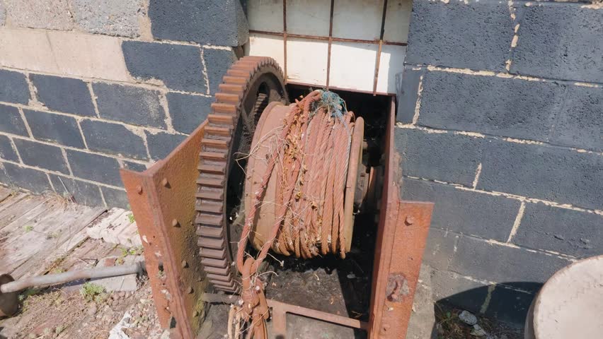 Rusted fishing boat winch with a large cog gear and coiled cable on Hastings beach, England. Camera pans above and then down, showing tangled netting around the rope.