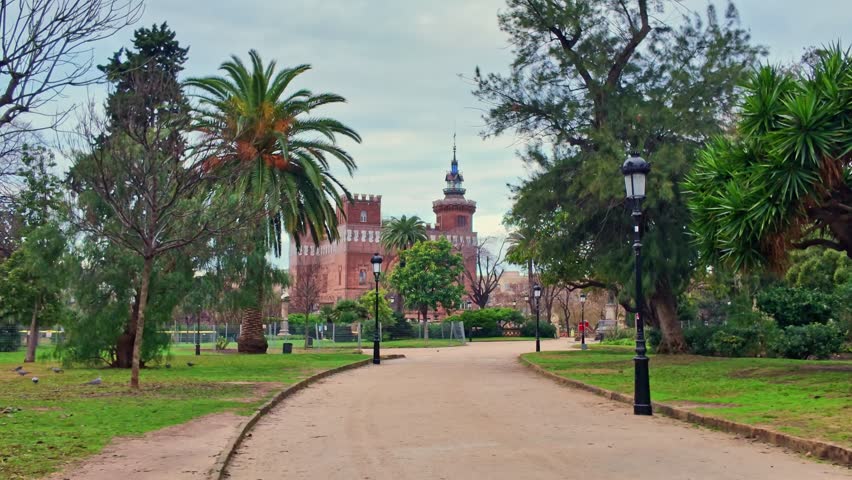 Forward view along a park path leading to Castell dels Tres Dragons in Barcelona, framed by palm trees and winter greenery.