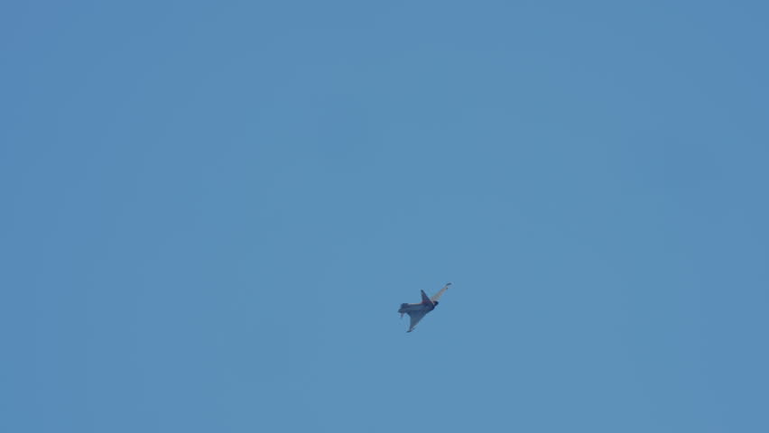 A military fighter jet performs an aerial maneuver, banking and flying across a clear blue sky in slow motion
