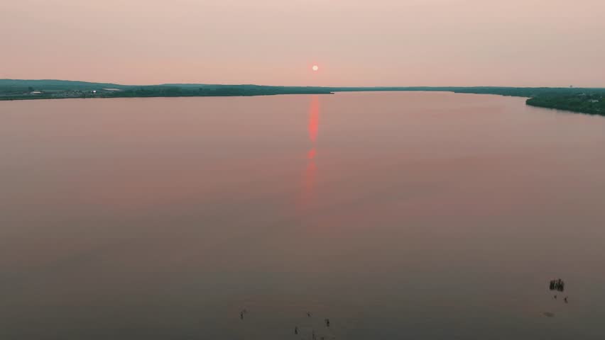 A lake wrapped by an undulating shoreline that extends on into the horizon as the sun hovers just above it.