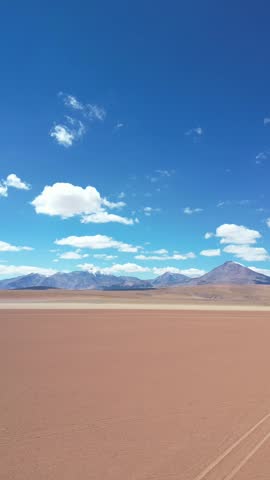 Retreating flight over red truck driving on desert road with vast sandy plains and volcanic mountain range under blue sky with scattered clouds. Atacama Desert, Chile