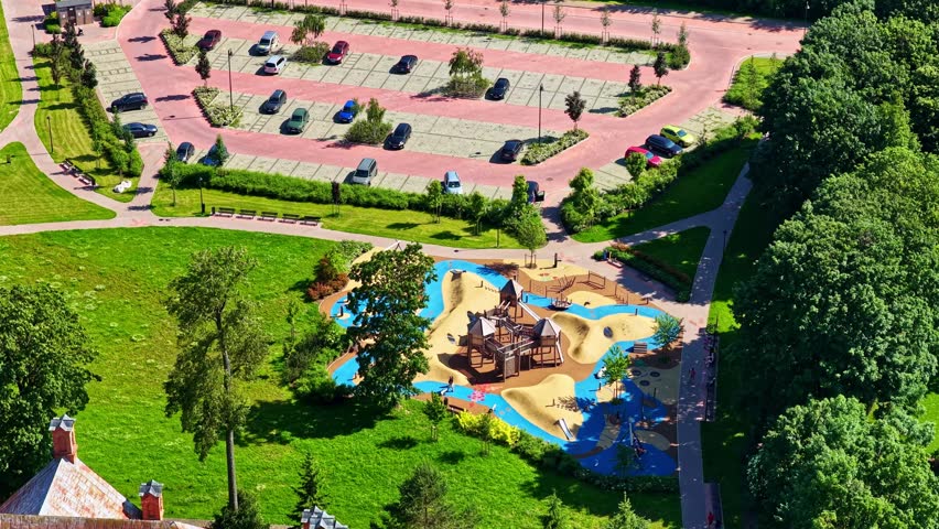 Aerial view of a modern playground with colorful flooring and a nearby parking lot surrounded by green trees in Riga, Latvia.