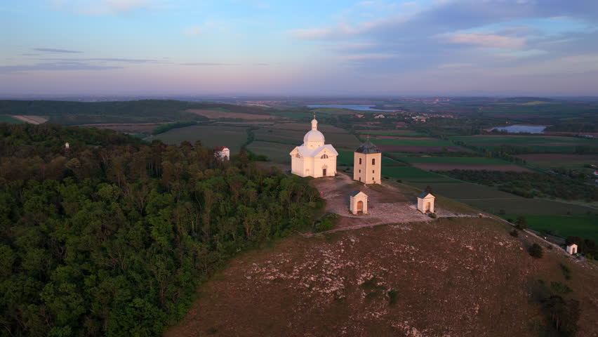 High-altitude aerial view of Holy Hill (Svaty Kopecek) with its Way of the Cross chapels and white baroque chapel above forests and vineyard plains at dusk, Mikulov, South Moravia, Czech Republic.