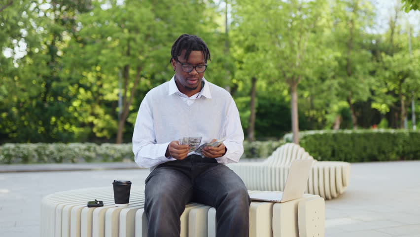 A happy young african american man with dreadlocks and glasses is holding cash and celebrating outdoors in a park setting