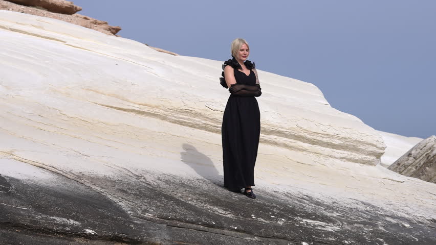 Blonde woman in a long black evening dress and gloves posing gracefully on white limestone rocks against a clear blue sky on a sunny day