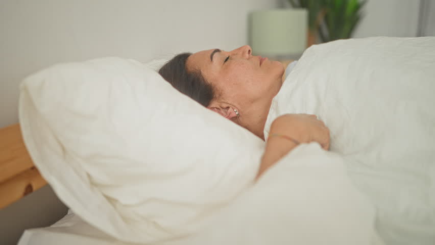 Woman smiling and hugging pillow in bedroom, lying on white duvet with hand showing ring and bracelet; serenity morning comfort.