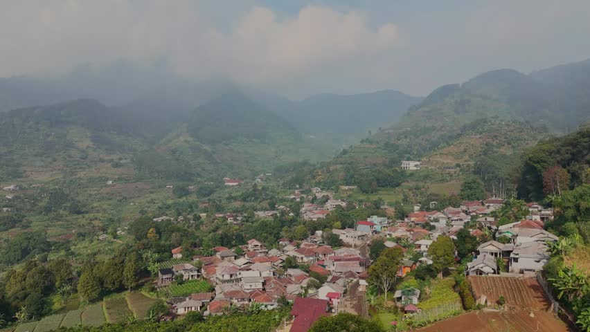 Aerial panoramic view of a rural settlement surrounded by steep forested hills and mist-covered mountain peaks in a tropical highland.