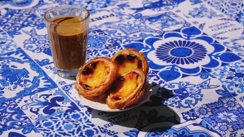Hand placing, adjusting and picking three pastel de nata egg tarts on white saucer. Traditional Portuguese pastry, glass of meia de leite coffee, blue and white tablecloth with azulejo motifs, blurred ackground, outdoor setting, high angle shot. Translation: best things in life are not things. Joy isnt found. It is made