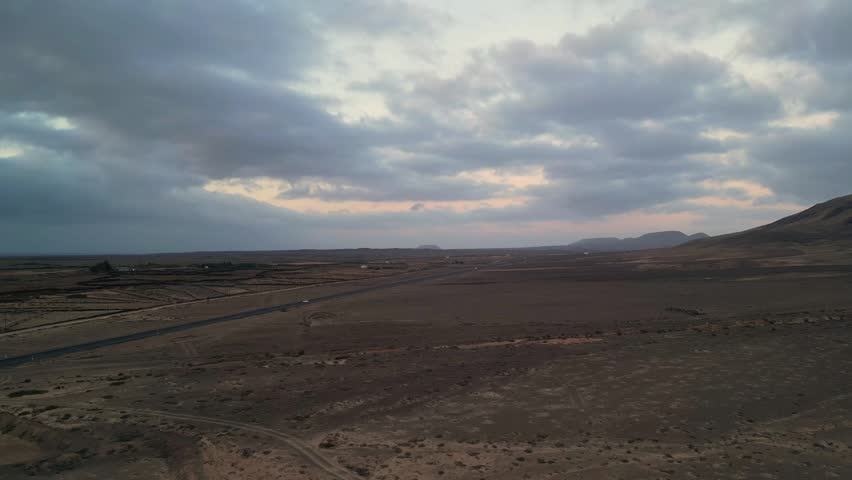 Aerial view of the arid desert landscape with a road stretching through the volcanic terrain under a cloudy sky in El Cotillo, Canary Islands, Spain.
