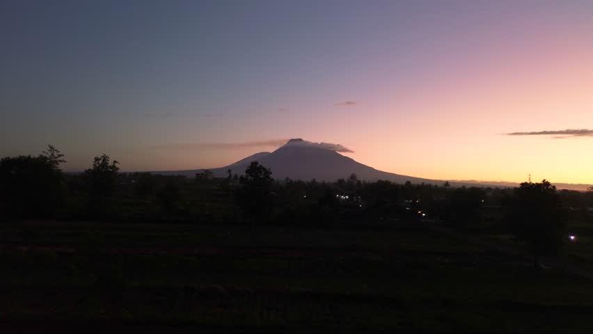 A stunning panoramic view of Mount Merapi from a distance, set against a dark pre-dawn sky. Aerial footage capturing the beauty of Mount Merapi while the sky is still dark.