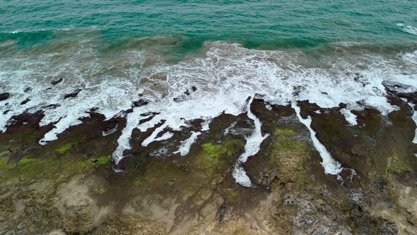 Aerial view of turquoise ocean waves crashing against the mossy rocky shoreline of Indonesia.