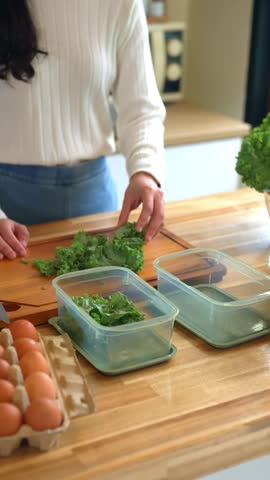 Young woman chopping fresh kale on a wooden cutting board with eggs and plastic containers, ideal for healthy eating and meal prep content