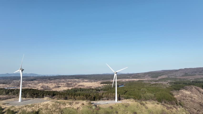 Aerial footage of the Nikaho Highlands South Observatory in Akita Prefecture, Japan. Captured on a clear spring afternoon, showcasing the snow-covered Mount Chokai and numerous wind turbines spread across the highlands. A unique landscape where renewable energy facilities blend with magnificent natural scenery.