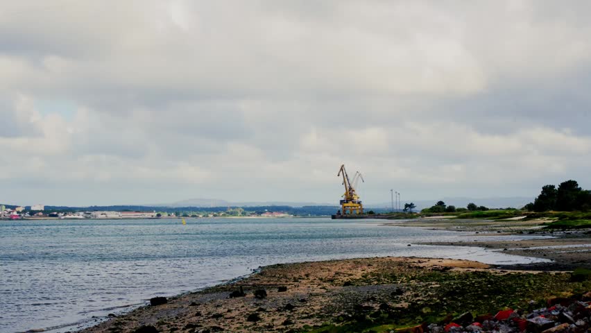 Coastal view of Firth of Clyde in Greenock Scotland. Calm water, rocky shoreline and industrial crane stand beneath cloudy sky along the estuary coastline