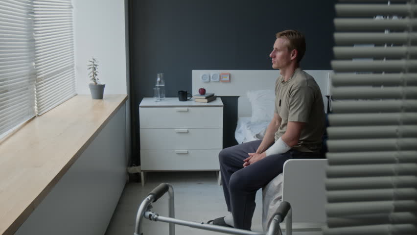 Tilt down shot of young adult Caucasian man with bandaged arm sitting on hospital bed near walker, looking out window. Video for healthcare, rehabilitation, and physical therapy promotions
