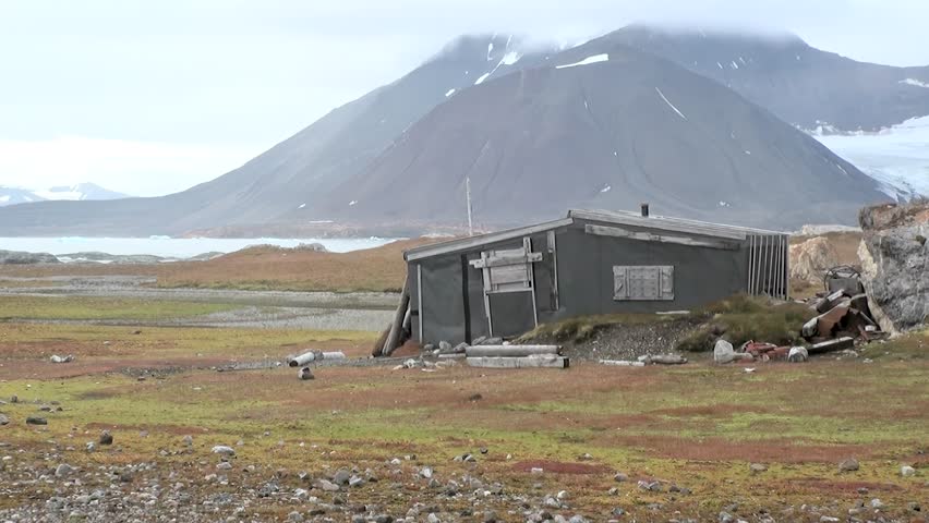 An ancient cabin rests on the vast tundra of Svalbard, Norway. Overcast skies loom above, creating a somber yet stunning Arctic panorama of mountains and sea. This remote structure endures the wild elements.