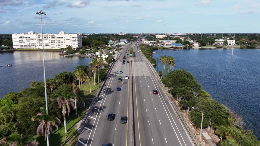 Divided causeway bridge carries cars beneath palm-lined median toward distant town, calm blue waterway flanking roadway on both sides near Merritt Island, Florida coast. Aerial drone wide shot