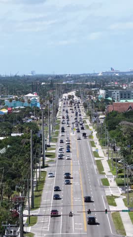 Dense traffic fills tree-lined coastal road as pedestrians cross marked crosswalks, distinctive dome-shaped building rising among beach homes in Cocoa Beach, Florida. Aerial drone vertical shot