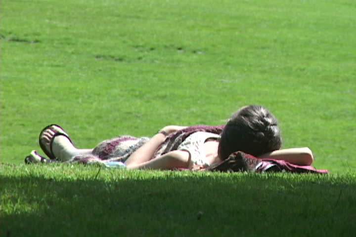 Woman lays relaxed in Portland Oregon park during sunny summer day. Dog runs in background.