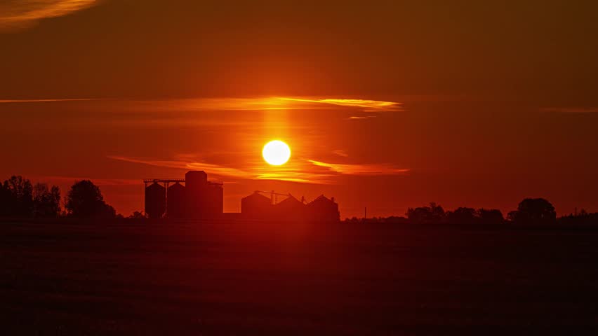 View of grain silos silhouetted against a vibrant orange sunset sky in the countryside of Latvia.