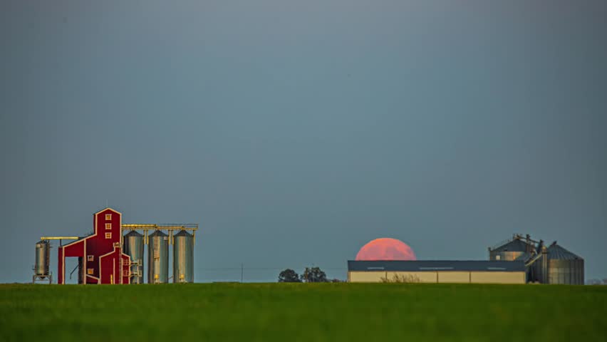 View of a large red moon rising behind farm buildings and metal grain silos in a rural landscape in Latvia.