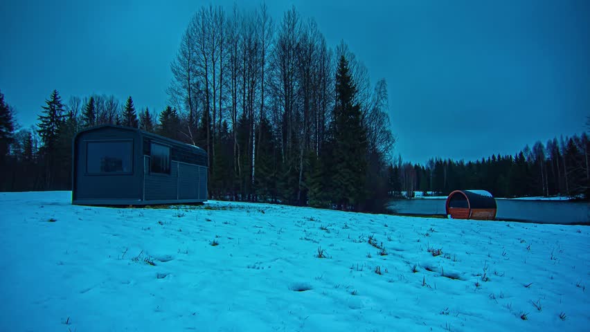 View of a modern cabin and a barrel sauna situated in a snow-covered field surrounded by a dense forest during the blue hour in Latvia.