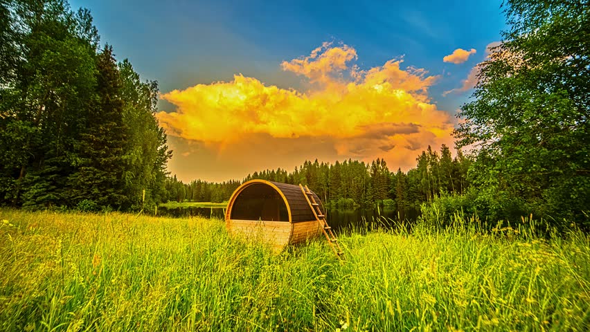 View of a wooden barrel sauna situated in a lush green meadow by a calm lake, framed by dense forest under a vibrant golden hour sky in Latvia.