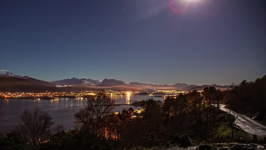 View of night cityscape of Alesund with illuminated buildings reflecting on the calm fjord waters under a starry sky with distant mountains, Alesund, More og Romsdal, Norway.
