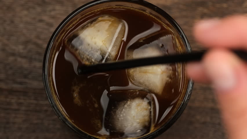 Iced coffee. Hand stirring coffee with ice in glass with black straw, close up