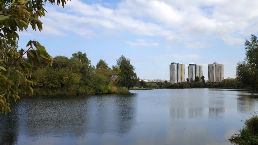 Bushes, willows and birches with branches bent over the water grow on the shore of the lake. Further on there are residential buildings and trees with yellowed leaves. Sunny autumn weather and blue sky with clouds