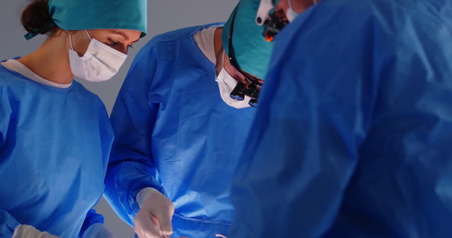 Surgeon group working, professional medical doctors surgical team performing surgery, doing operation, procedure on patient, wearing surgical gown, cap, face mask, eye loupe binocular, treat injuries