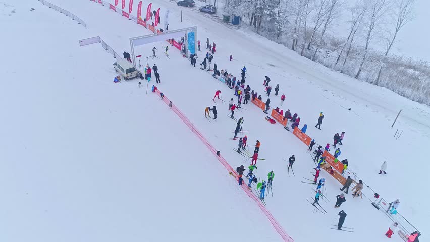 Aerial view of cross country skiing competition with athletes and spectators gathered at the start line in a snowy landscape in Latvia.
