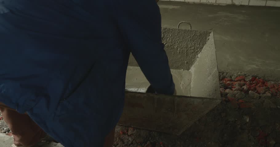 Professional builder in workwear taking liquid concrete with a bucket from a container and pouring it on the floor, leveling the surface of a new building during renovation work.