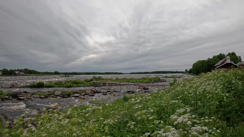4K static view of Kukkolankoski rapids in Finland with wildflowers, green grass, traditional fishing structures, and rushing river beneath dramatic overcast skies.