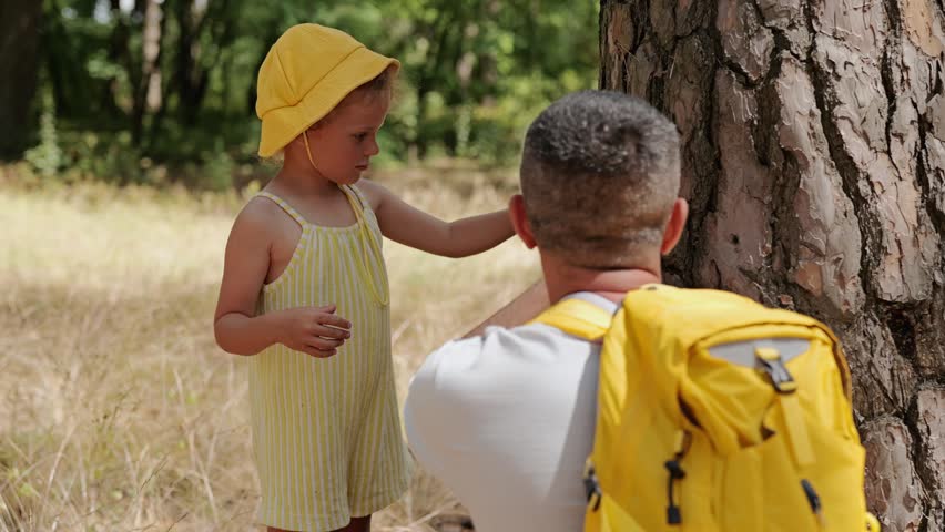 A child in a yellow hat gives a snack to an adult while standing near a tree in a forest. They enjoy a break together in the warm weather