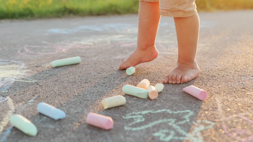 A child stands barefoot on the sidewalk surrounded by colorful chalk pieces. Chalk drawings cover the ground as the sun lights up the scene