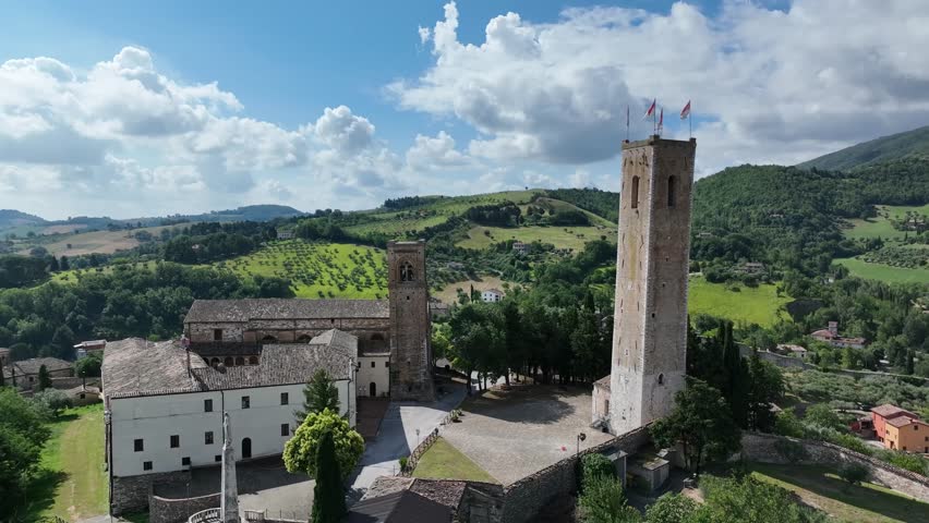 Visitors walk around a historic site with tall towers and a large open space. The site is surrounded by green hills and trees. Clouds cover the blue sky.
