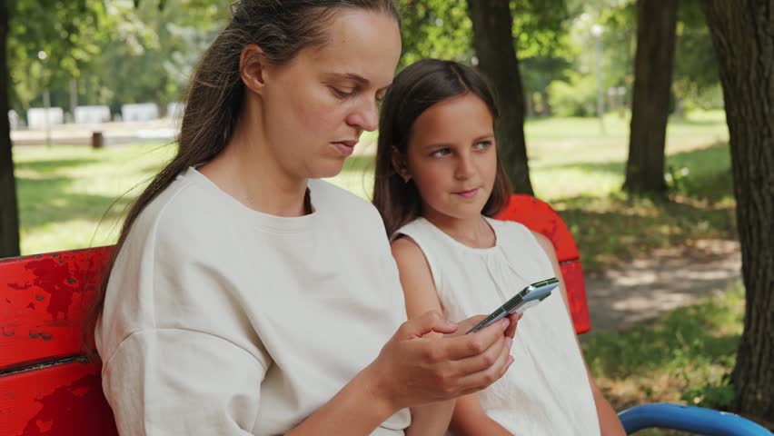 A mother and daughter enjoy time together in a park. They sit on a bench and look at a smartphone. Trees surround them on a sunny day