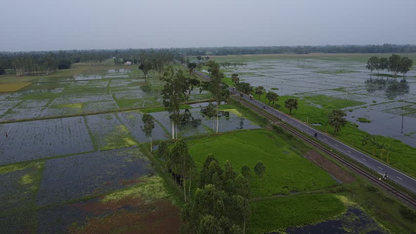 Aerial view of lush green flooded rice fields alongside a rural asphalt road and railway track with a motorized rickshaw traveling through countryside drone perspective looking down at vast