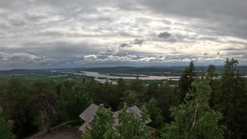 Breathtaking 4K panorama from Aavasaksa hill over the Tornio River valley, with pine trees, a wooden roof, and the Finland Sweden border beneath dramatic cloudy skies.