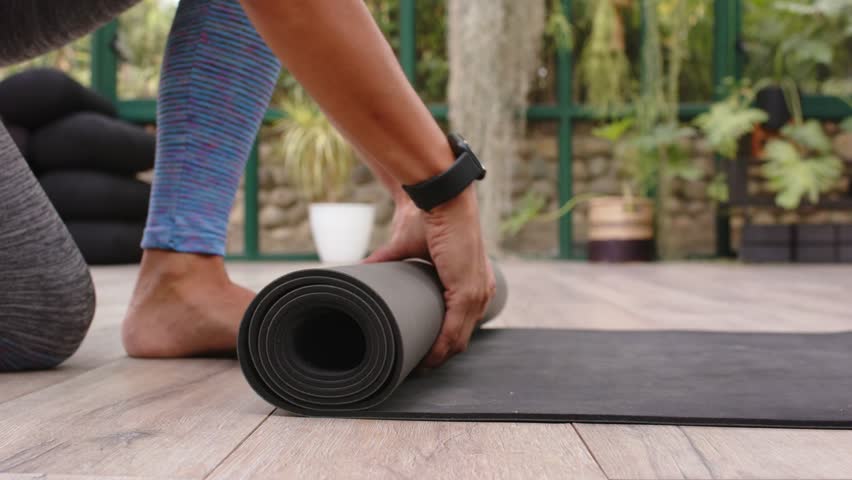 Woman reaching forward and pulling rolled black yoga mat, unrolling and smoothing for yoga practice. Conservatory, sunroom, daylight, plants, cushions, planks, fitness
