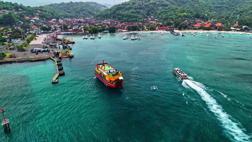 Aerial view of a ferry and speed boat navigating the turquoise waters near a coastal town with lush green hills and traditional buildings in Bali, Indonesia.