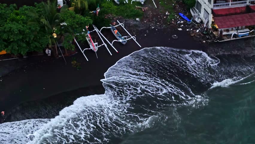 Aerial view of traditional outrigger boats resting on a black sand beach with waves crashing against the shore in Bali, Indonesia.
